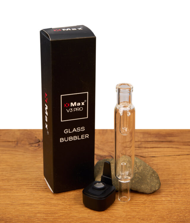 XMax V4 Pro Glass Bubbler neben schwarzer Verpackung. Transparenter Wasserfilter und magnetischer Adapter mit Metallsieb auf Holztisch. Auch mit V3 Pro kompatibel.