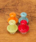 Vier Black Leaf Carb Cap Spinner aus buntem Glas (Orange, Blau, Grün, Rot) in der Draufsicht auf einer Holzoberfläche arrangiert.