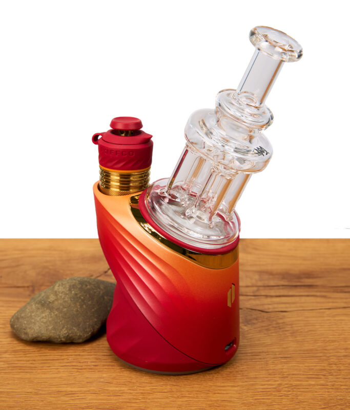 Das Puffco Peak Quadra Top von MJ Arsenal einsatzbereit montiert auf einem rot-orangefarbenen Vaporizer neben einem kleinen Naturstein.