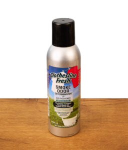 Eine silberne Sprühdose des Smoke Odor Spray Clothesline Fresh Lufterfrischers mit schwarzer Kappe auf einer Holzoberfläche; das Label zeigt frische Wäsche auf einer Wäscheleine.