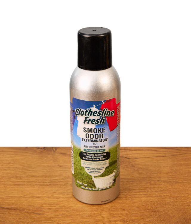 Eine silberne Sprühdose des Smoke Odor Spray Clothesline Fresh Lufterfrischers mit schwarzer Kappe auf einer Holzoberfläche; das Label zeigt frische Wäsche auf einer Wäscheleine.