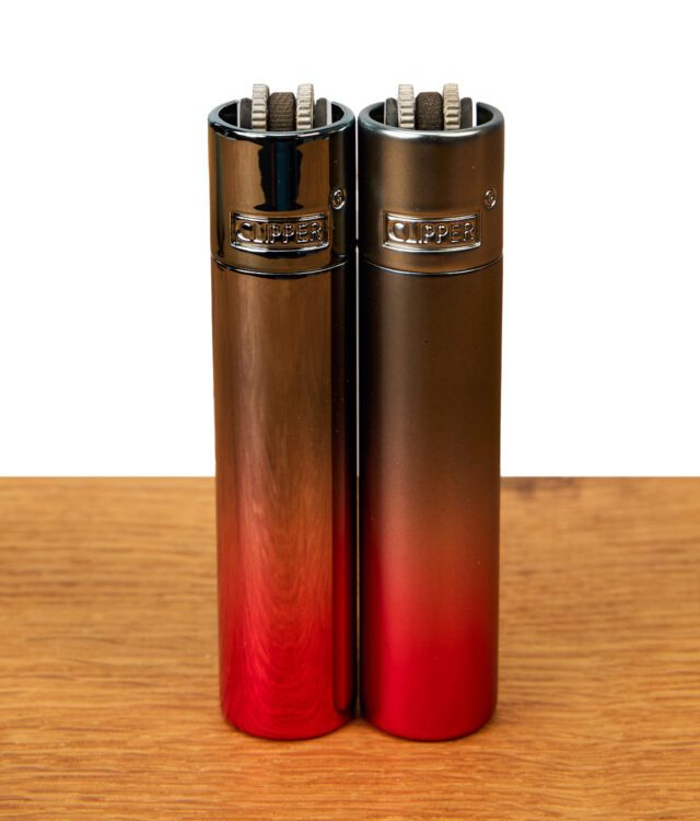 Zwei Clipper Metall Demon Gradient Feuerzeuge im direkten Vergleich nebeneinander auf Holz: Links die hochglänzende (Icy-Optik) und rechts die matte Variante des edlen Metall-Lighters mit rot-schwarzem Farbverlauf.