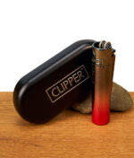 Das glänzende Clipper Metall Demon Gradient Sturmfeuerzeug präsentiert neben der klassischen schwarzen Clipper Metallbox auf einer Holzoberfläche mit kleiner Steindekoration.