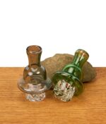 Zwei Spinner Carb Caps für Quarzbanger in den Farben Grau und Grün, dekorativ arrangiert vor einem Naturstein auf einer Holzoberfläche.