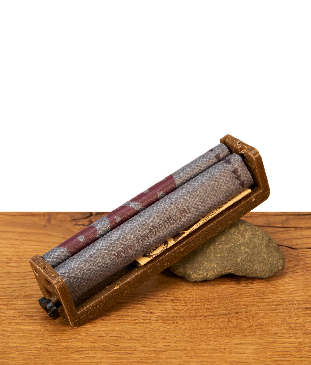 raw-eco-2-way-roller-drehmaschine-king-size-110mm-jpg RAW Eco 2-Way Roller Drehmaschine King Size 110mm aus Hanfplastik mit bedrucktem Rollband, gestützt auf einen Stein auf einem Holztisch.