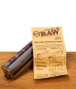 Einstellbare RAW Eco 2-Way Roller Drehmaschine King Size 110mm aus Hanfplastik, liegend neben der aufgeschlagenen Bedienungsanleitung auf Holz.