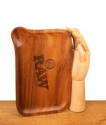 Hochkantes RAW Rolling Tray aus Holz. Hell gemaserte Drehunterlage mit Einfüll-Ecke neben einer hölzernen Gliederhand zum Größenvergleich.
