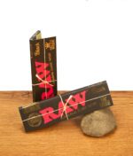 Zwei Päckchen RAW Black King Size Wide Paper. Die schwarzen Heftchen mit rotem Logo und Goldprägung liegen auf Holz und Stein.