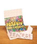 Aufgeklapptes Juicy Jay's Happy Birthday Paper auf Holz. Buntes Innendesign mit entnehmbaren, bedruckten Kuchen-Blättchen.