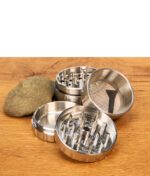 Geöffnete Kammern des Steely Dan 2.0 Edelstahl Grinders neben einem Stein auf Holztisch, Fokus auf das feine Pollensieb.