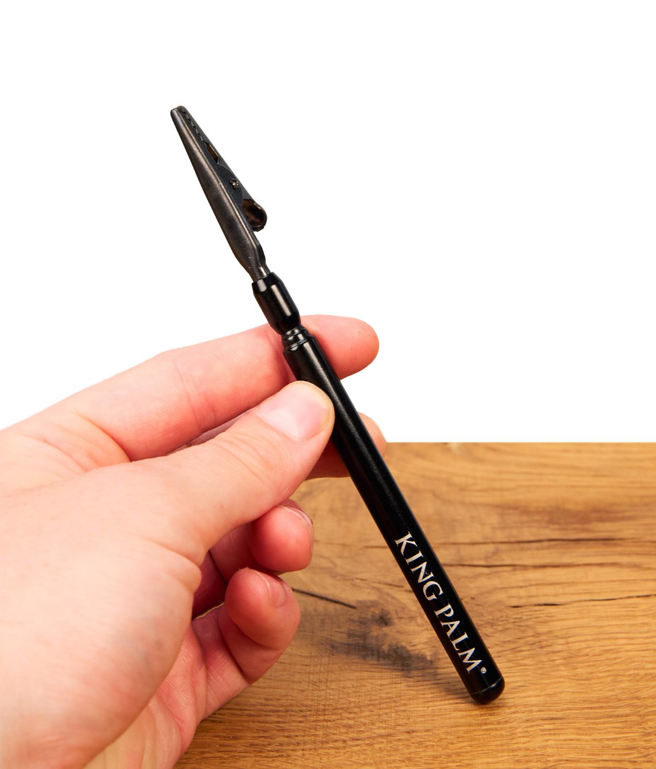 king-palm-blunt-clip-jpg King Palm Blunt Clip aus schwarzem Metall mit weißem Schriftzug, in der Hand gehalten über einer Holzoberfläche.
