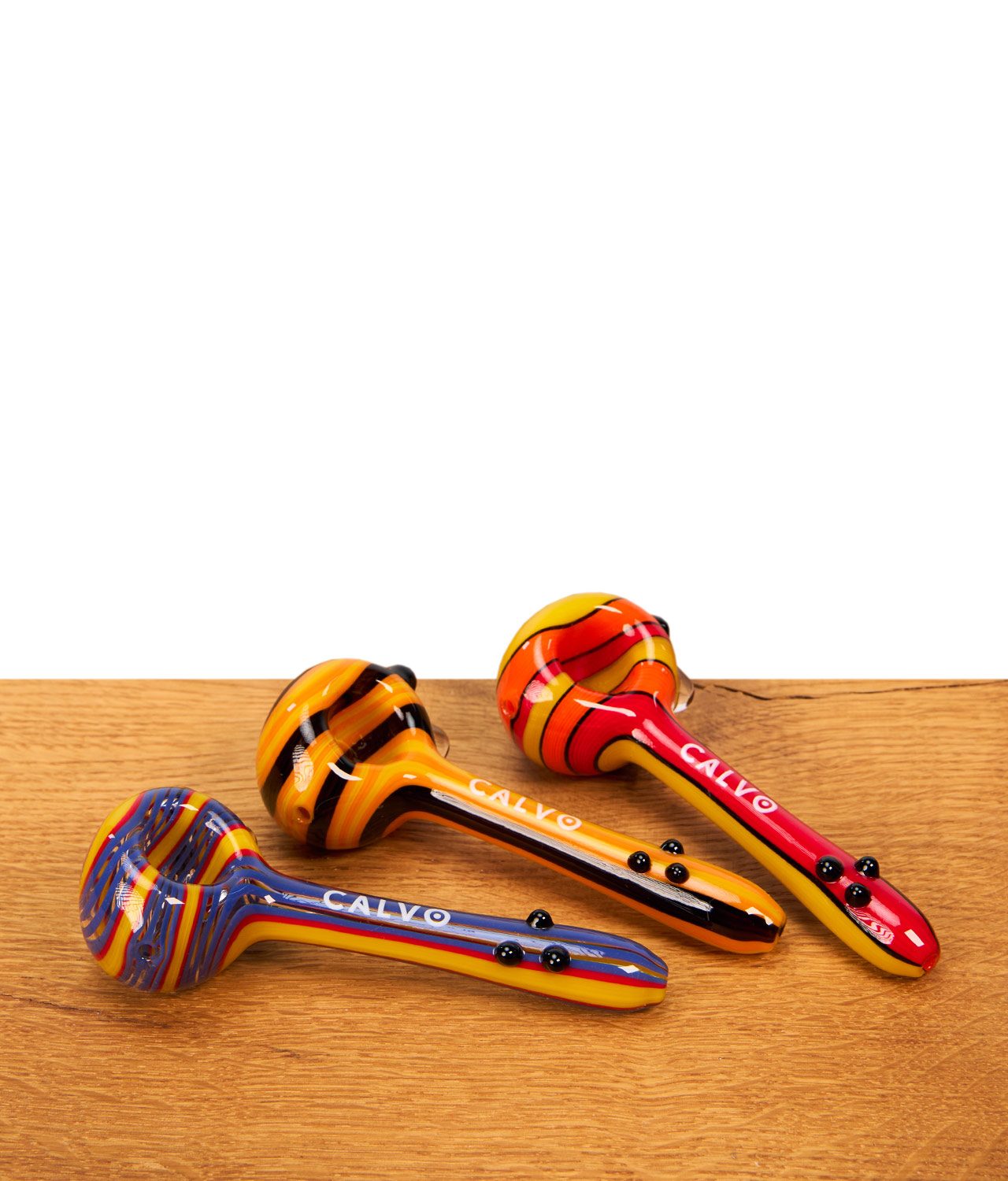 b1-calvo-unique-spoon-pipe-gruppenbild-jpg Drei farbenfrohe Calvo Unique Spoon Pipe Glaspfeifen mit Streifenmuster nebeneinander auf Holzoberfläche.