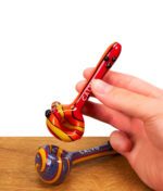 Eine Hand hält die rot-gelbe Calvo Unique Spoon Pipe Glaspfeife über einer Holzoberfläche.