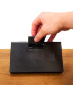 Eine Hand öffnet das Batteriefach auf der Rückseite der BL Scale Digitalwaage im Notizbuch-Design auf Holz.