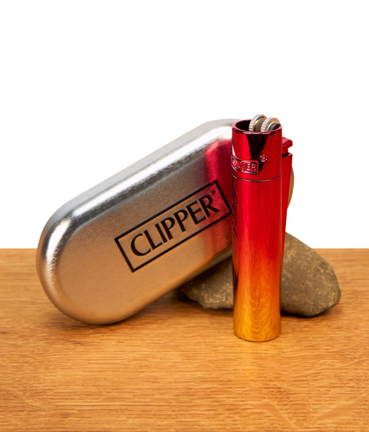 clipper-metall-sunset-gradient-jpg Das schimmernde Clipper Metall Sunset Gradient Feuerzeug steht aufrecht neben seiner geschlossenen, ovalen Präsentationsbox aus Metall auf einem Holztisch vor weißem Hintergrund.