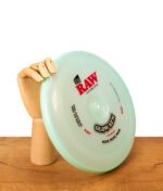 RAW Frisbee Cyan Glow SZN Special Edition, aufrecht gehalten von einer hölzernen Gliederhand auf einem Holztisch.