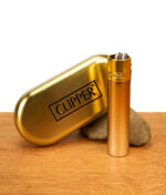 Mattes Clipper Metall Gradient Elegance Gold Feuerzeug stehend neben geprägter Metalldose und Stein auf Holz.