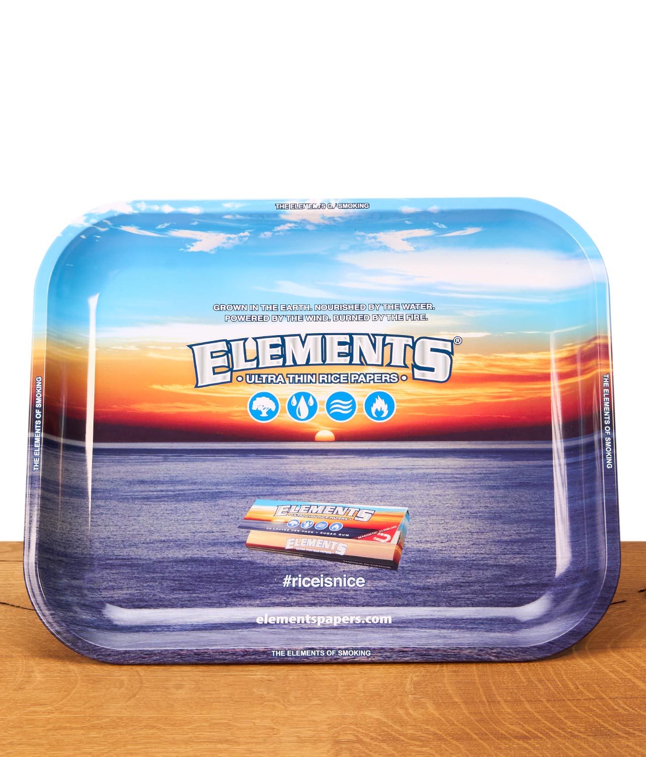 Elements-Rolling-Tray-Gross-Blue Rechteckiges Elements Rolling Tray aus Metall auf einem Holztisch. Die Drehunterlage zeigt ein Ozean-Sonnenuntergang-Motiv mit Markenlogo.