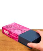 Eine Hand zieht die edle Box aus der pinken, illustrierten Verpackung des GRAV Ellipse Aschenbechers auf einem Holztisch.