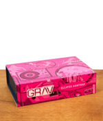 Geschlossene, pinkfarben illustrierte Produktverpackung des GRAV Ellipse Glas-Aschenbechers auf einer Holzoberfläche.