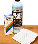 Randy's Black Label Cleaner Flasche neben Randy's Swipes Reinigungstüchern und deren Verpackung auf einer Holzplatte.