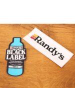 Zwei farbige Randy's Marken-Sticker, einer im Design der Black Label Reiniger-Flasche, auf einer strukturierten Holzoberfläche.