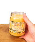 Geöffnete Smoke Odor Bubbly Mimosa Kerze aus Parasoy-Wachs im Glas, von einer Hand über einer Holzplatte gehalten.