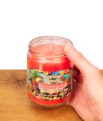 Hand hält die offene Smoke Odor Duftkerze Tropical Fiesta auf einem Holztisch. Zeigt das rote Wachs im Glasgefäß mit buntem Etikett.