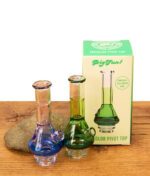 Blauer und grüner BigFun! Tricolor Pivot Top Glasaufsatz neben der grünen Originalverpackung und Stein auf Holz.